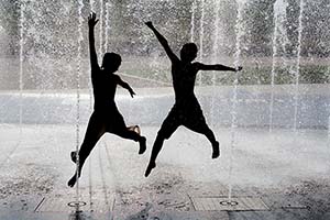 silhouette of a pair of young men trying to stay cool by jumping through water sprayed up by fountains in an urban setting