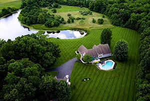 Overhead view of a beautifully landscaped field of grass and trees with a massive, fantastic pond surrounding a huge home and an inviting swimming pool where the yellow dog can play