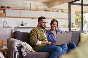 young couple of color smiling on the couch staring at a laptop going over a mortgage application, assessing their chances that a lender will let them have the money they need to own a home