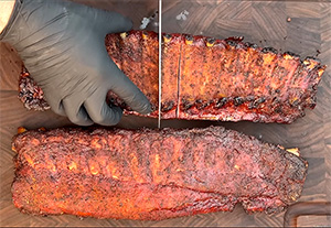 a gloved hand holds down on a rack of ribs seasoned and smoked to perfection on a wood-grained cutting board as a knife slices up the ribs into individual servings with one for the yellow dog
