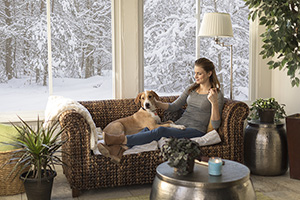 big windows in the room show a snowy wintery scene of cold while a woman seated on a couch near the windows with dog seems warm and comfortable in her warmer older home upgraded and winterized
