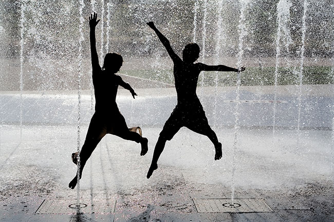 silhouette of a pair of young men trying to stay cool by jumping through water sprayed up by fountains in an urban setting