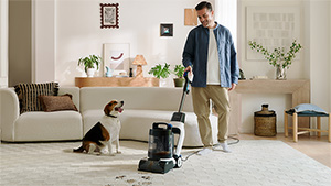 smiling young guy runs a carpet cleaner over muddy paw prints on the white living room rug as the dog watches with his (dog) tongue hanging out